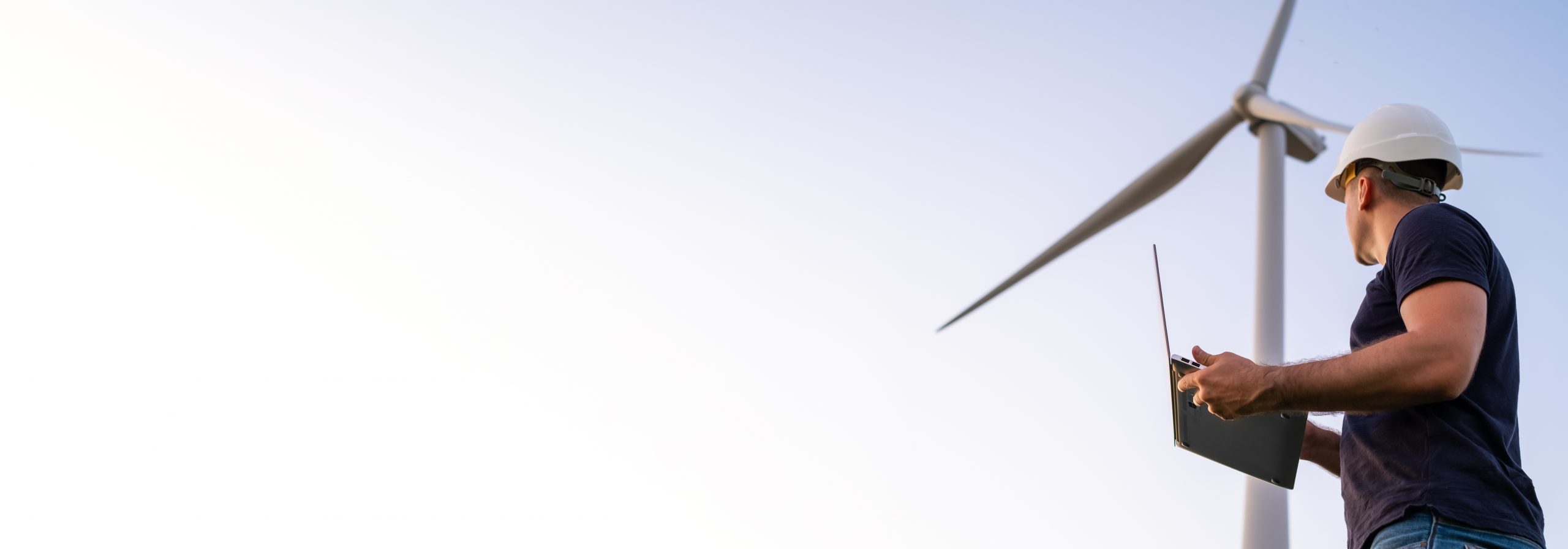 Eine Person, die einen Schutzhelm trägt und einen Laptop in der Hand hält, steht in der Nähe einer großen Windturbine und inspiziert oder überwacht diese unter einem klaren, hellen Himmel.
