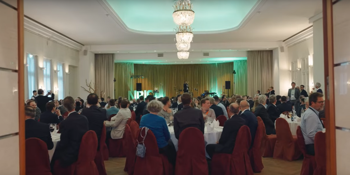 Eine formelle Veranstaltung in einem großen, eleganten Bankettsaal mit Kronleuchtern. Die formell gekleideten Gäste sitzen an runden, mit weißen Tischtüchern bedeckten Tischen und blicken auf eine Bühne, auf der Vadība und die beleuchteten Buchstaben NPI zu sehen sind.