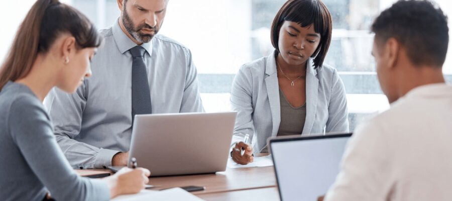 Vier Personen in formeller Kleidung, die konzentriert an einem Tisch mit Laptops und Notizbüchern arbeiten. Die Atmosphäre ist professionell und fokussiert.
