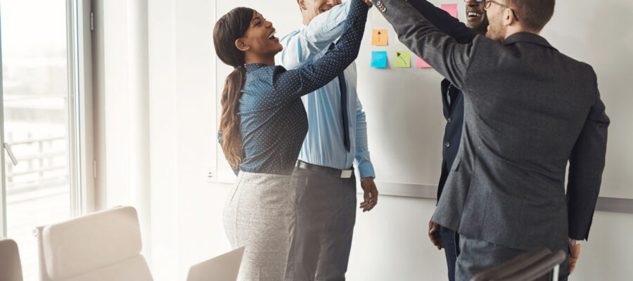 Drei Männer und eine Frau stehen in einem Besprechungsraum vor einer Wandtafel, an dieser hängen Notizzettel. Die Vier scheinen scheinen ein Erfolgserlebniss gehabt zu haben und geben sich ein "High Five".