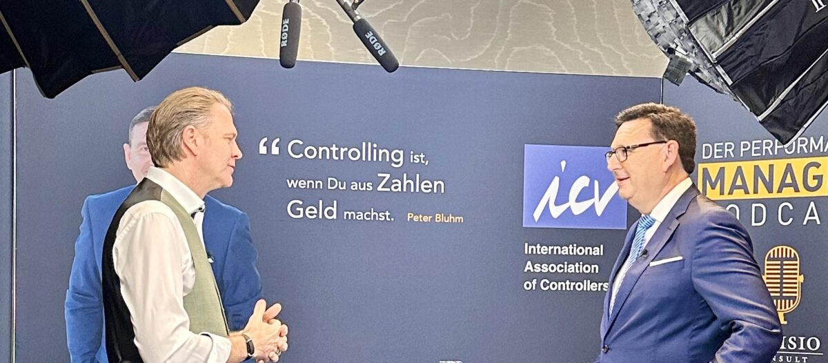 Zwei Männer in Geschäftskleidung diskutieren über Vadība vor einer Kulisse mit der Aufschrift "Controlling ist, wenn Du aus Zahlen Geld machst". Über ihnen hängen Mikrofone und Studiolampen.