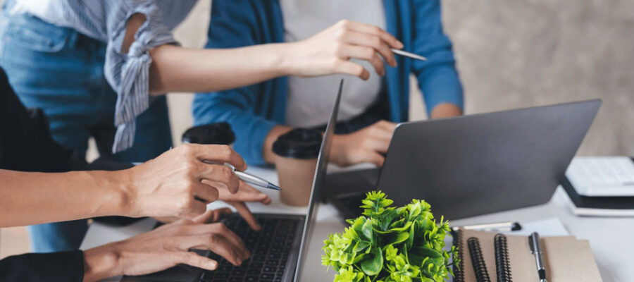 Eine Gruppe von Personen, die gemeinsam an einem Tisch arbeiten. Sie nutzen Laptops und zeigen mit Stiften auf die Bildschirme, während sie offensichtlich über etwas diskutieren. Auf dem Tisch befinden sich außerdem Notizbücher, Stifte, zwei Kaffeebecher und eine kleine grüne Pflanze. Die Szene vermittelt eine dynamische und kooperative Arbeitsatmosphäre.