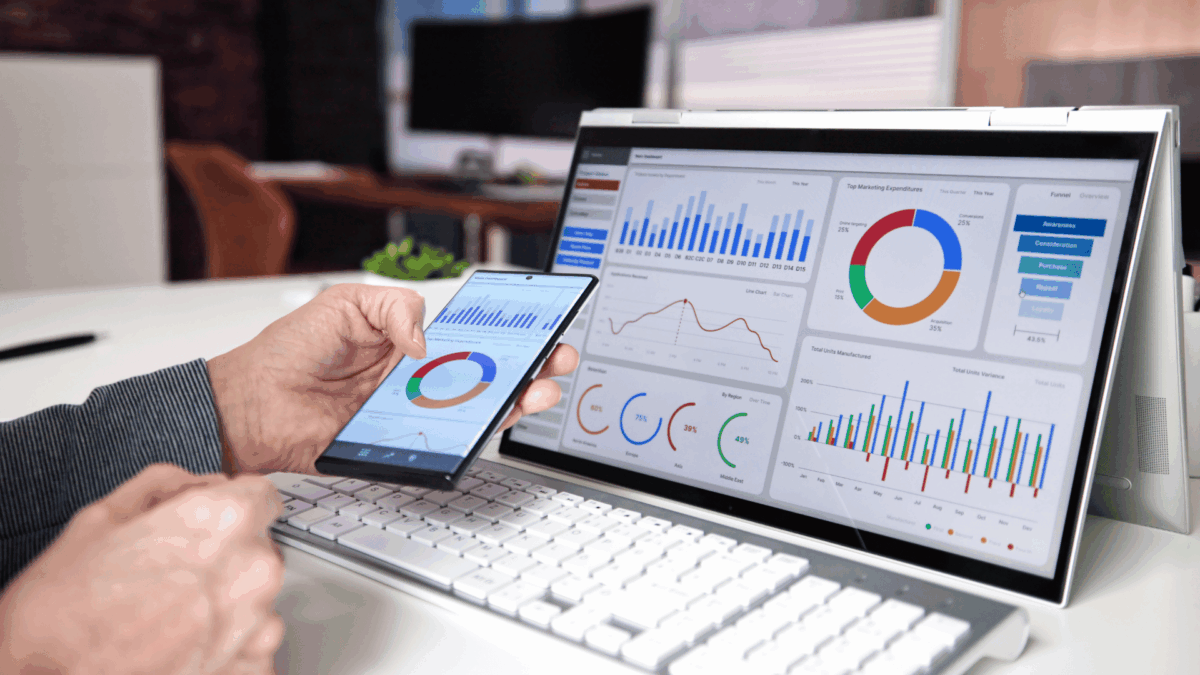 Person analysiert Marketing- und Vertriebsdaten auf Smartphone und Laptop, beide Geräte zeigen bunte Diagramme und Kennzahlen in einem modernen Büro.