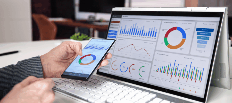 Person analysiert Marketing- und Vertriebsdaten auf Smartphone und Laptop, beide Geräte zeigen bunte Diagramme und Kennzahlen in einem modernen Büro.