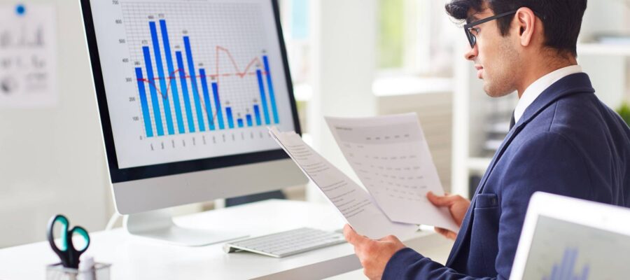 Abweichungsanalyse Titelbild - Ein Mann im Anzug analysiert Daten vor einem Computer und schaut sich dazu Papierunterlagen an.