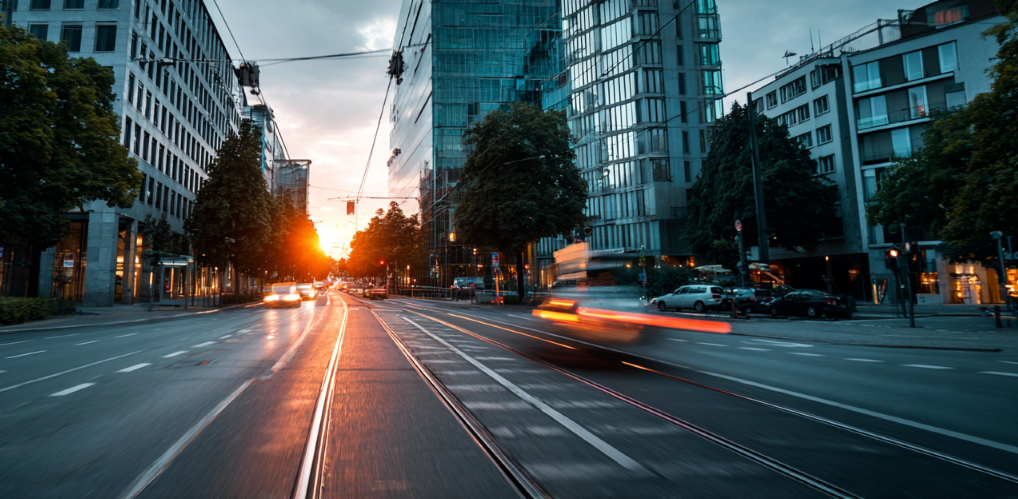 Stadtansicht einer modernen deutschen Stadt mit Infrastrukturelementen wie Straßen, Bürogebäuden, Autos. Der Fokus liegt auf dem Zusammenspiel von digitaler und analoger Infrastruktur. Weiches Licht und dezente Bewegungsunschärfe durch vorbeifahrende Autos. Neutrale Farbtöne, leichte urbane Atmosphäre. Keine Menschen im Bild – nur Raum und Funktion.