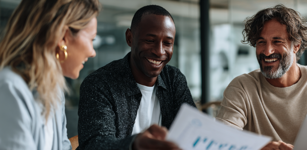 Drei Personen in einem entspannten Besprechungsraum unterhalten sich über ausgedruckte Berichte und Laptop-Bildschirme, eine Person zeigt auf ein Diagramm. Der Raum ist hell, mit großen Fenstern, natürlichem Licht und moderner, schlichter Einrichtung. Mimik und Körperhaltung vermitteln Zusammenarbeit, Vertrauen und Engagement. Die Szene wirkt authentisch und nicht gestellt.