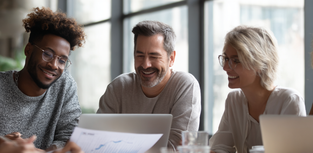 Drei Personen in einem lockeren Besprechungssetting diskutieren über ausgedruckte Berichte und Laptop-Bildschirme, eine Person zeigt auf ein Diagramm. Der Raum ist hell und modern eingerichtet, mit großen Fenstern und weichem, natürlichem Licht. Gesichtsausdrücke und Körperhaltung vermitteln Kooperation, Vertrauen und Engagement. Die Szene wirkt authentisch und nicht inszeniert.