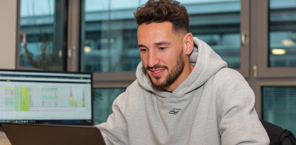 Unternehmensplanung bei einem Fußball Sportverein. In einem hellen Büro sitzt eine Person und schaut konzentriert auf seinen Laptop. Leicht verschwommen sieht man im Hntergrund auf einem Bildschirm eine Spieltagsanalyse mit Spieltags-Kennzahlen. Im Hintergrund sind große Fensterscheiben. Durch die Fenster sieht man im Hintergrund ein Nachwuchsleistungszentrum.
