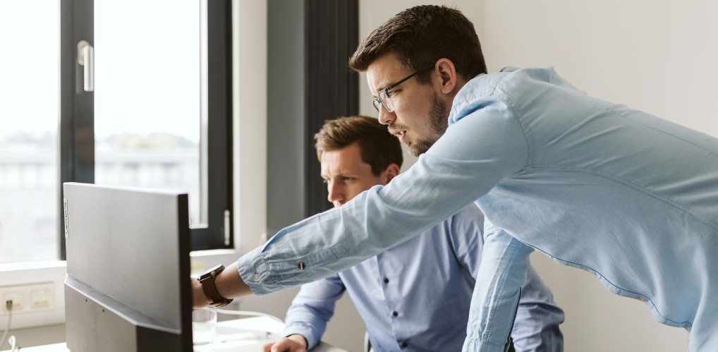 Das Bild zeigt zwei Kollegen von TD Trusted Decisions in einem hellen Büro. Der eine sitzt am Arbeitsplatz. Der andere steht vor dem Schreibtisch und zeigt auf den Bildschirn vor ihnen.